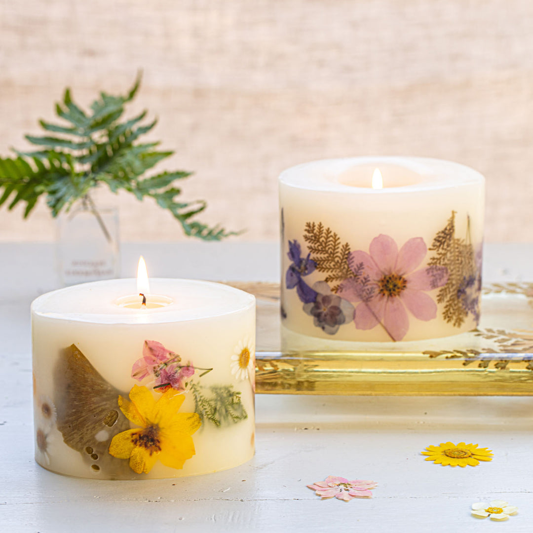 Two floral candles, one lit, with dried flowers and scattered petals on a soft backdrop for a delicate, serene scene.