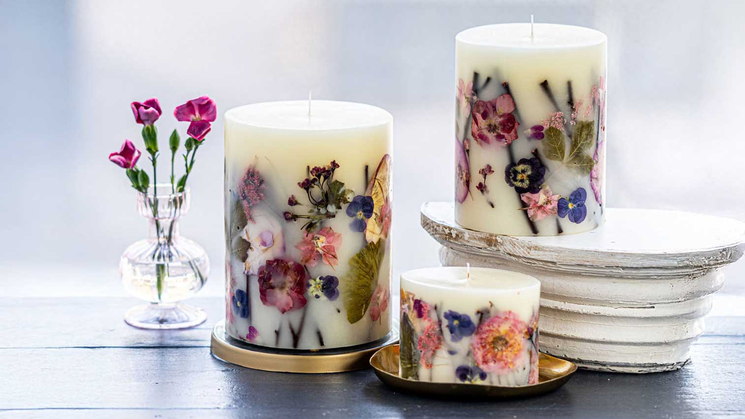 Decorative candles and diffusers with dried flowers on a rustic wooden table, set against a bright background.