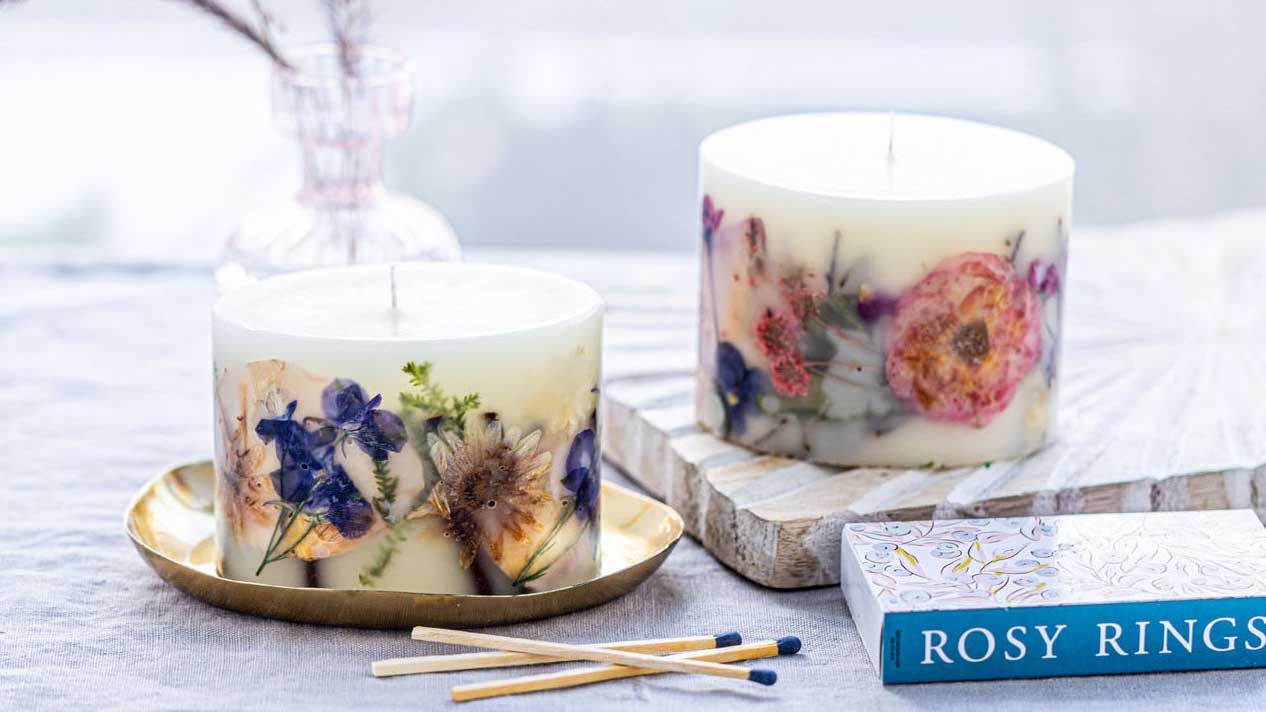 Two decorative candles featuring pressed flowers, with matchsticks and a book titled Rosy Rings nearby.