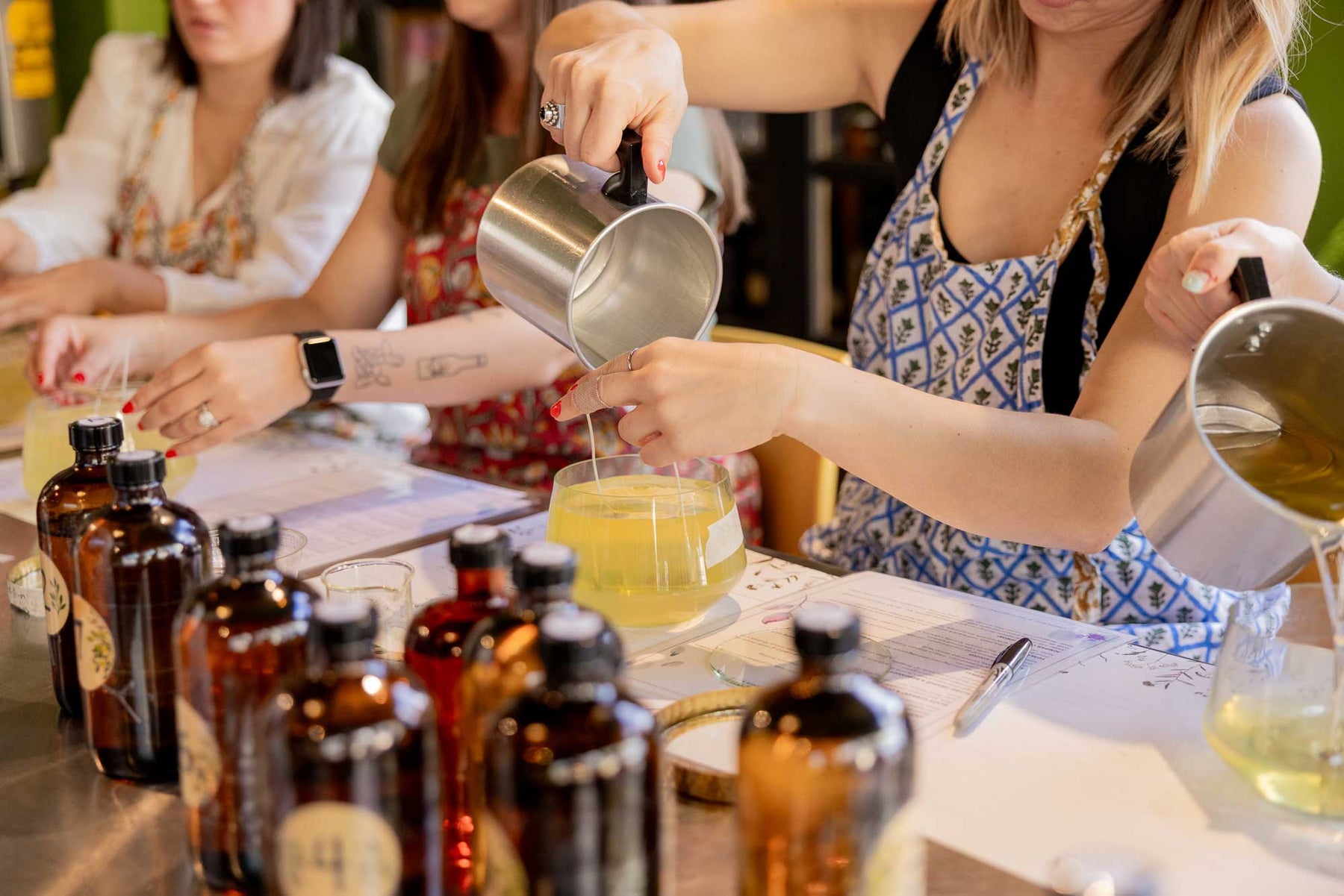 Workshop participants pour liquids into containers, surrounded by amber bottles and ingredients on the table.