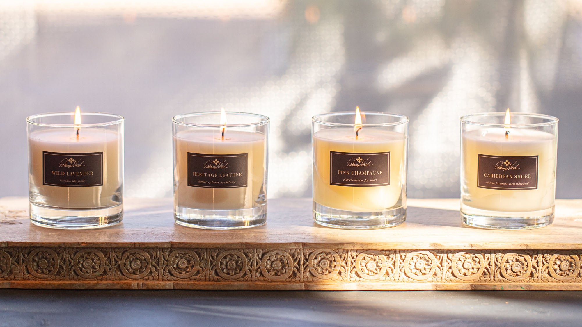 Four lit candles in glass jars labeled with unique scents, displayed on a carved wooden tray in soft natural light.
