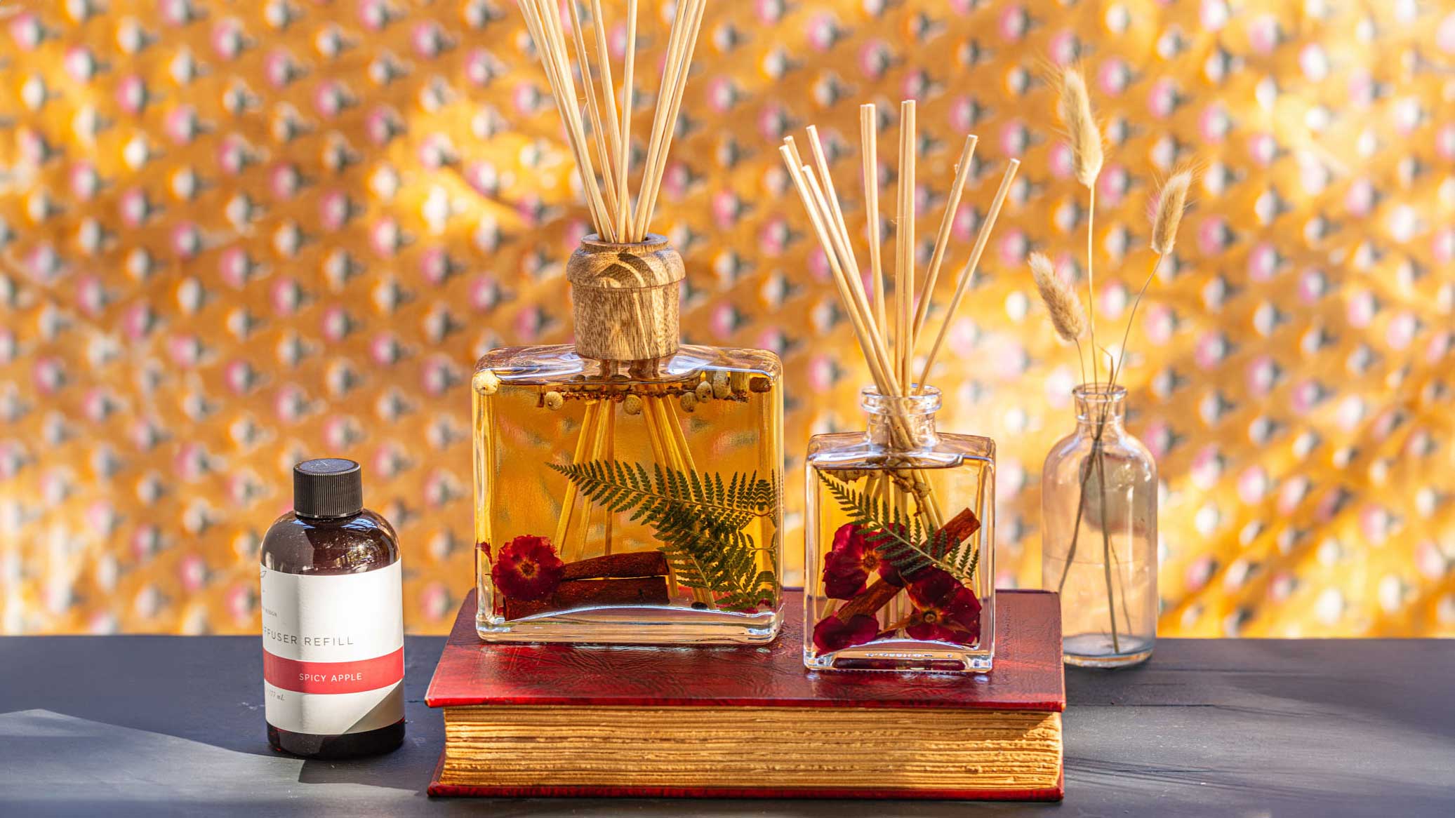 Two floral reed diffusers and a Spicy Apple refill bottle arranged on a red book.