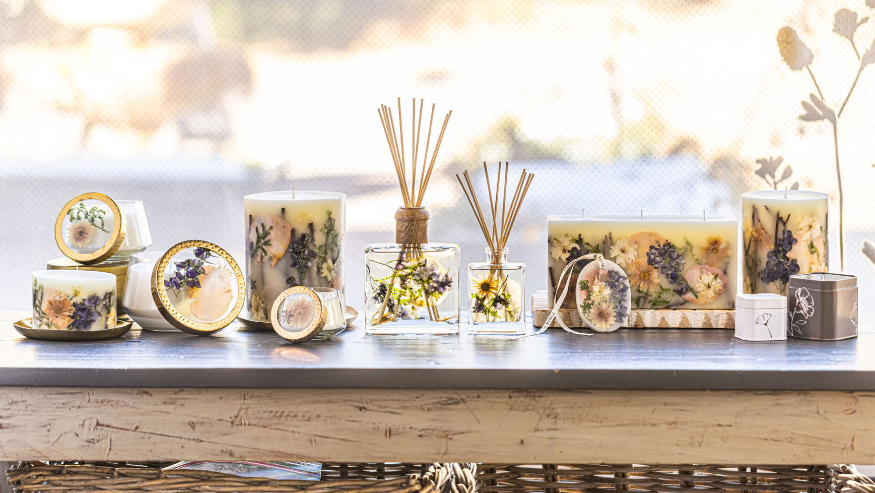 Decorative candles and diffusers with dried flowers displayed on a wooden table against a bright, sunlit background.