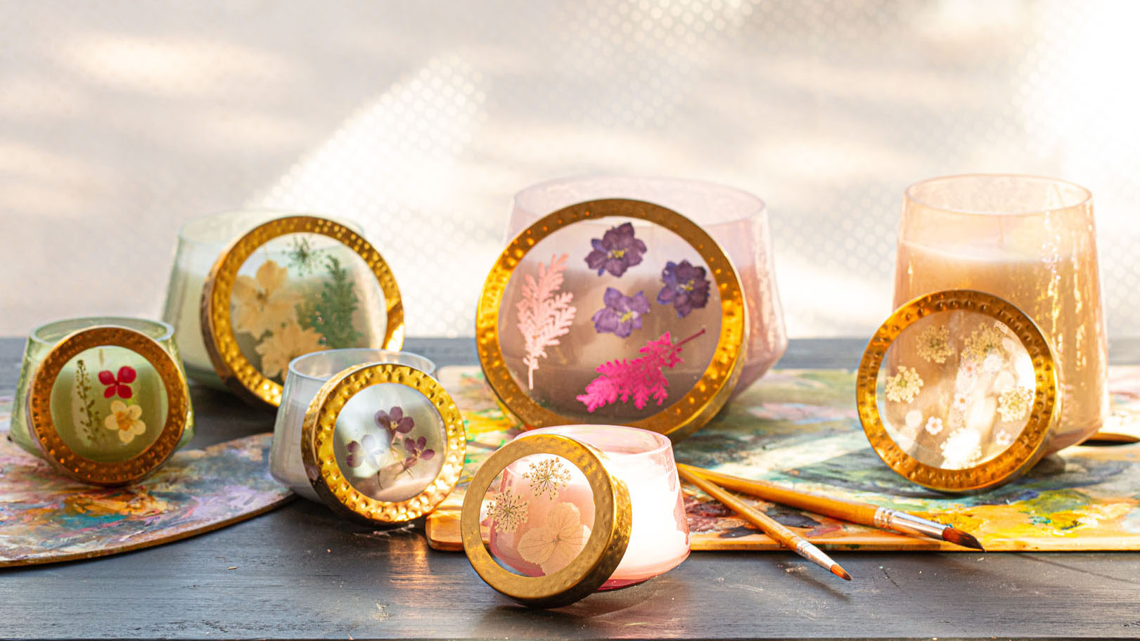 Pressed flower candles in gold-topped glass holders arranged on a vibrant artist’s palette for a creative display.