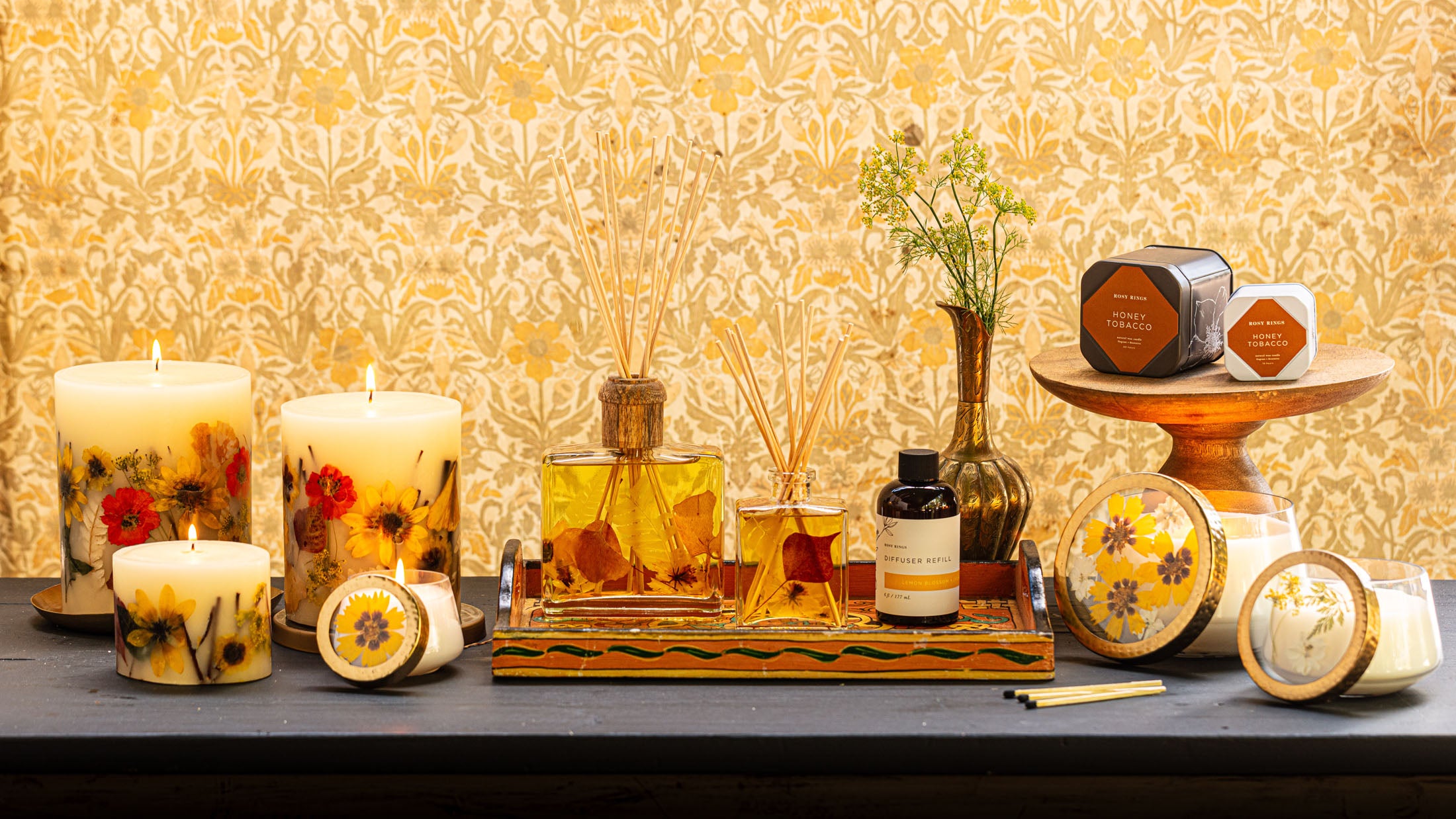 Floral candles diffusers and fragrance containers artfully arranged on a table with a patterned golden backdrop.