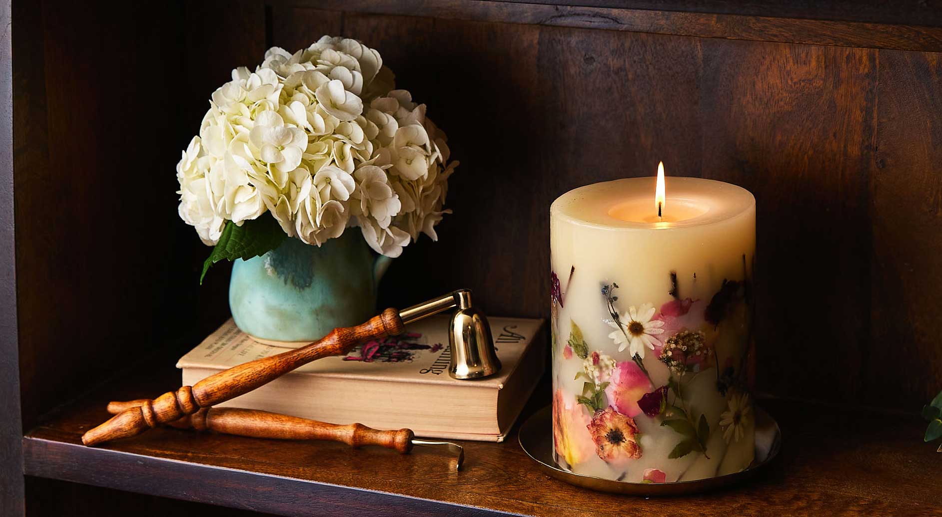 A lit floral candle sits beside a small bouquet of white hydrangeas in a turquoise vase, resting on a wooden shelf.