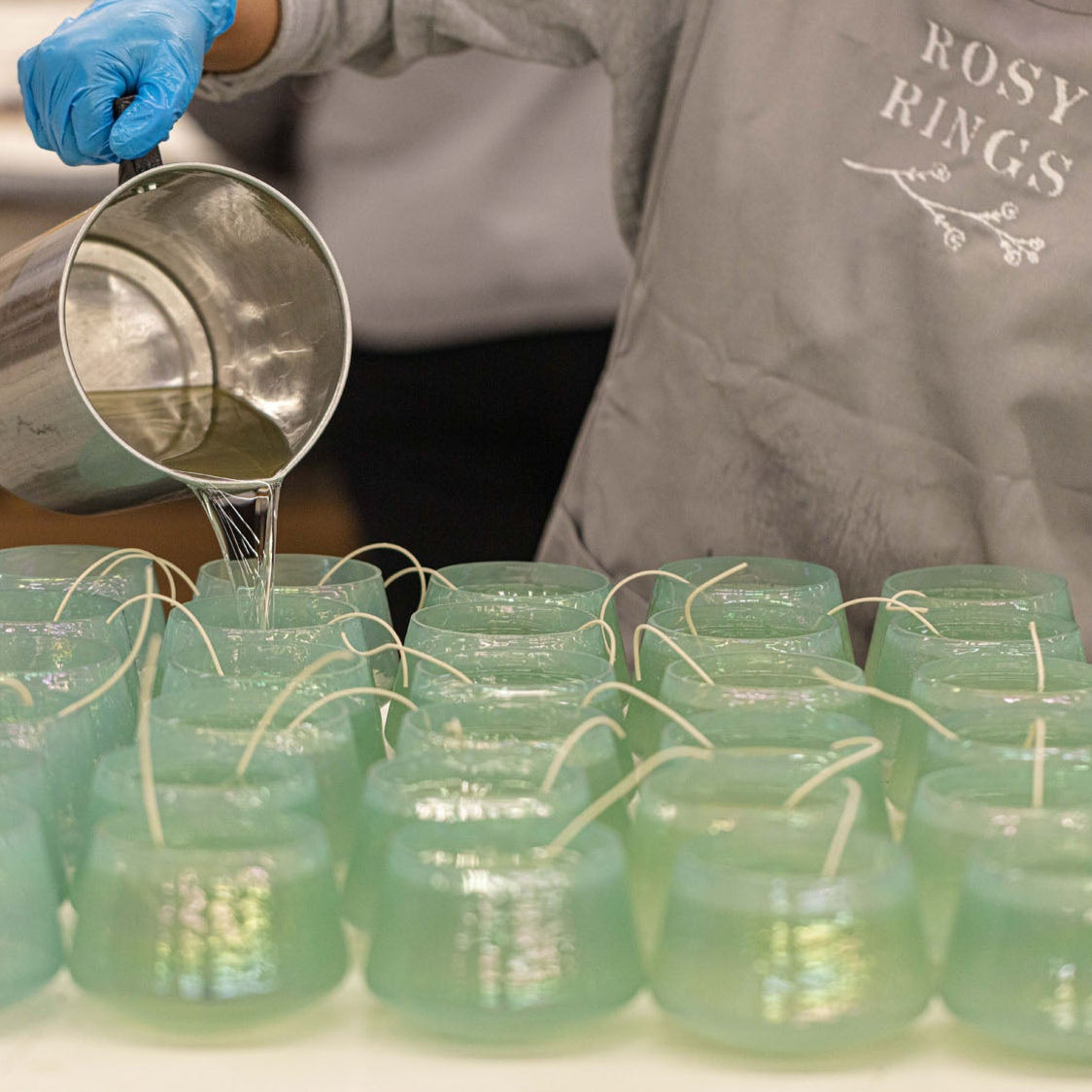 Person pouring liquid into small green jars with a strainer, wearing a 'Rosy Rings' apron.