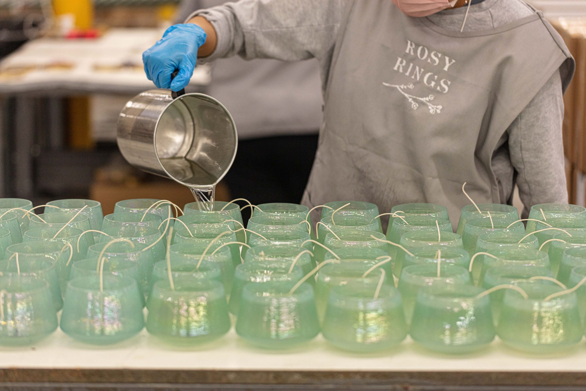 Gloved worker pours wax into mint-green candle molds with wicks in a Rosy Rings production area.
